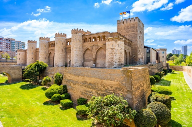 A sand-colored castle with turrets and walls, surrounded by green grass