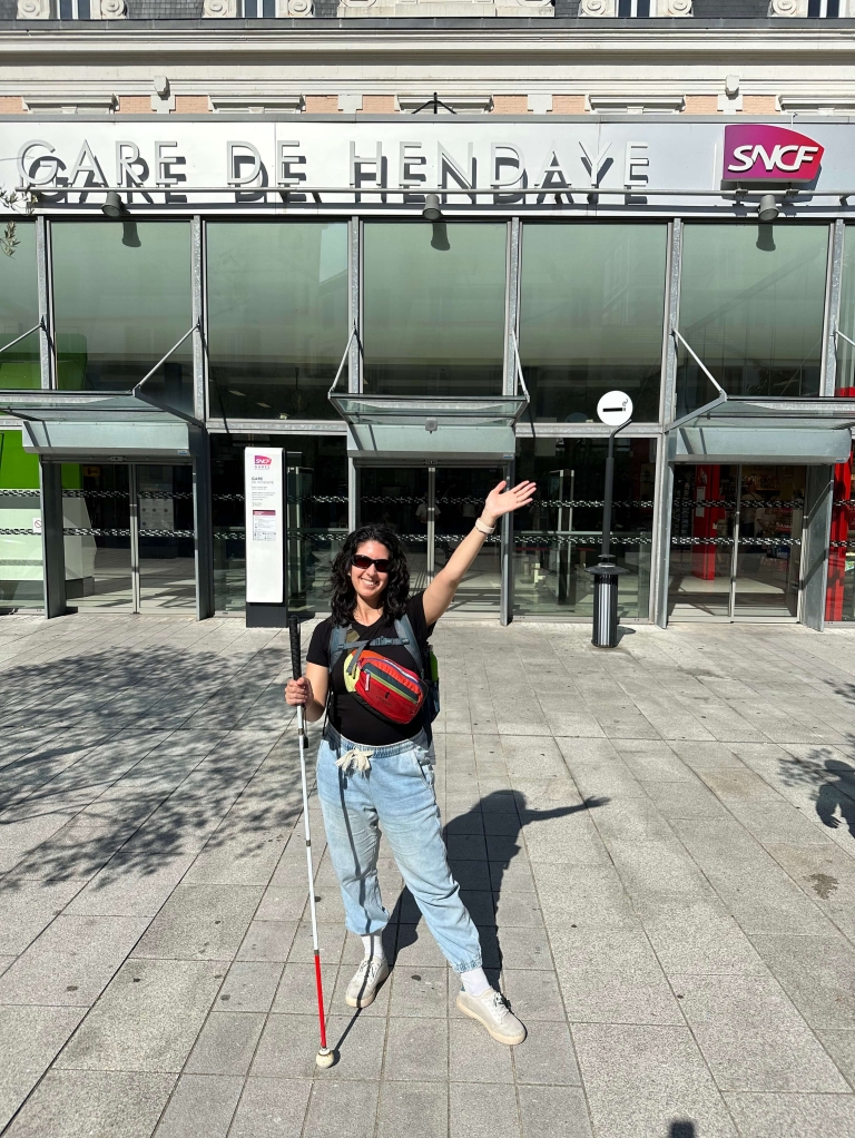 A woman holding a white and red cane strikes a pose outside a train station that says "Gare de Hendaye"