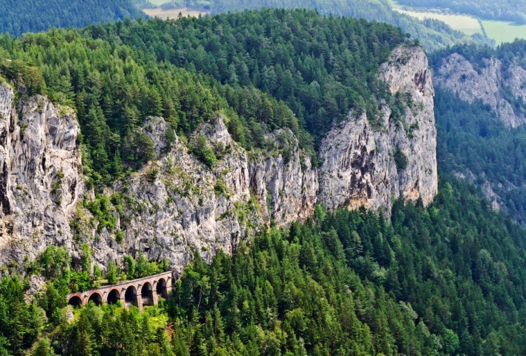 A dramatic, forested cliff, with a train bridge in the lower right of the frame