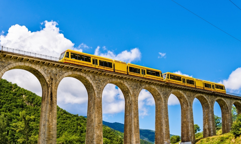 A yellow train on top of a wide bridge connecting lush hills